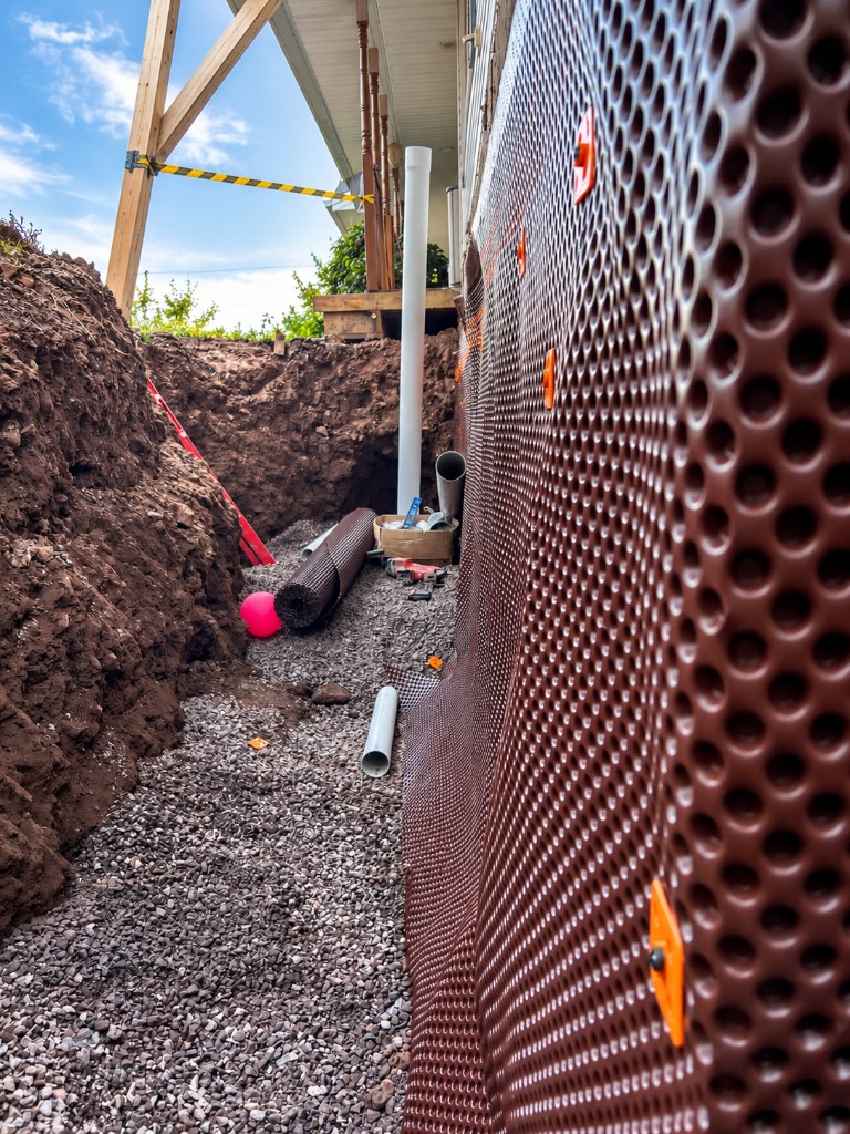Dimpled foundation membrane and gravel drain tile installed in a trench along a house, with the porch held up on posts