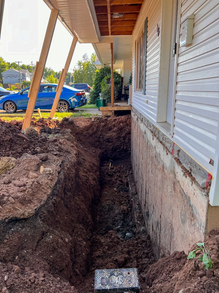 A trench dug along a house foundation for drain tile repair, with the porch held up on wooden posts