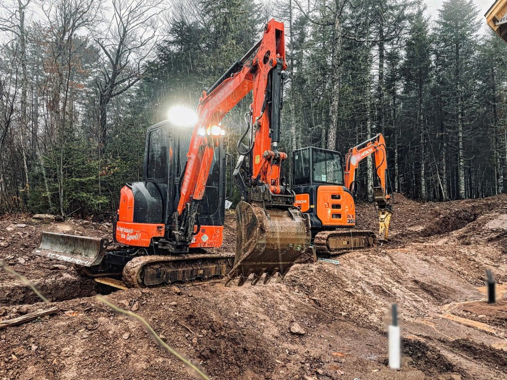 Two orange Choreguys Hitachi excavators on a muddy job site in the woods