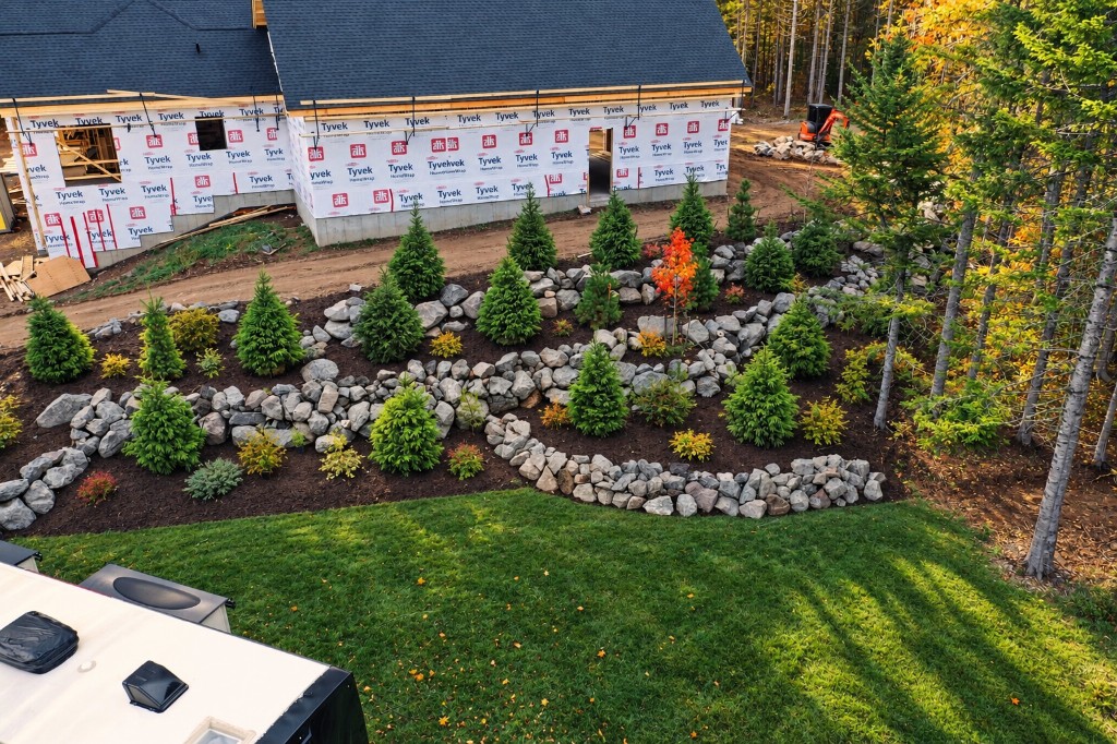 An elevated view of curved natural rock walls and young evergreens in front of a house under construction, with a lawn in the foreground