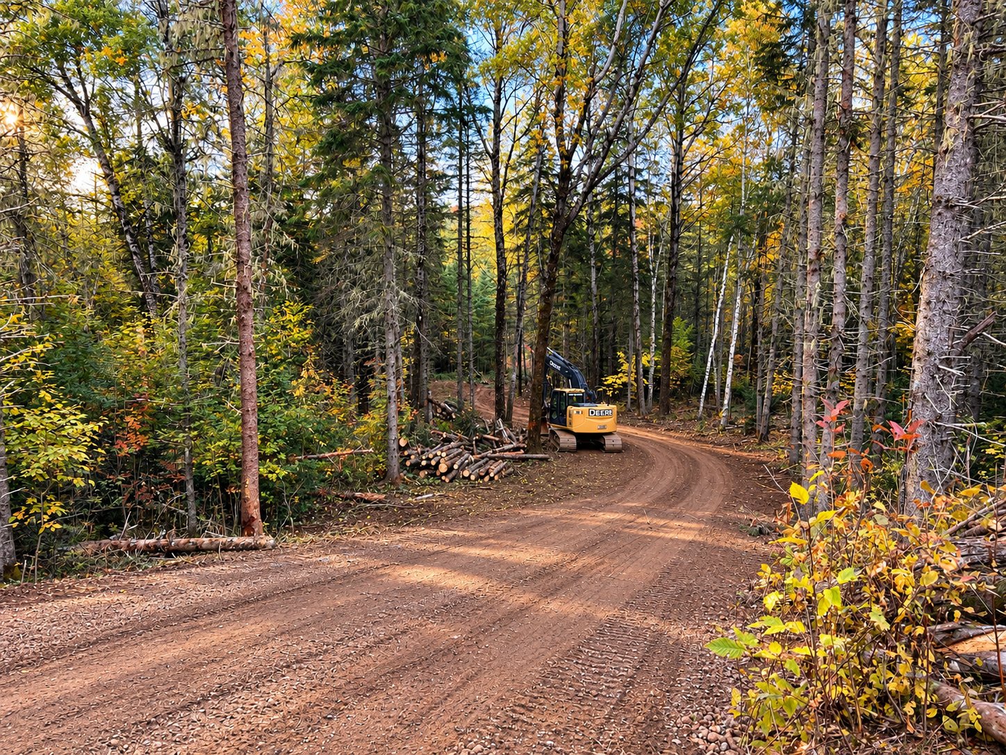 A new gravel road curving through a fall woodlot, with a John Deere excavator and a pile of cut logs beside it