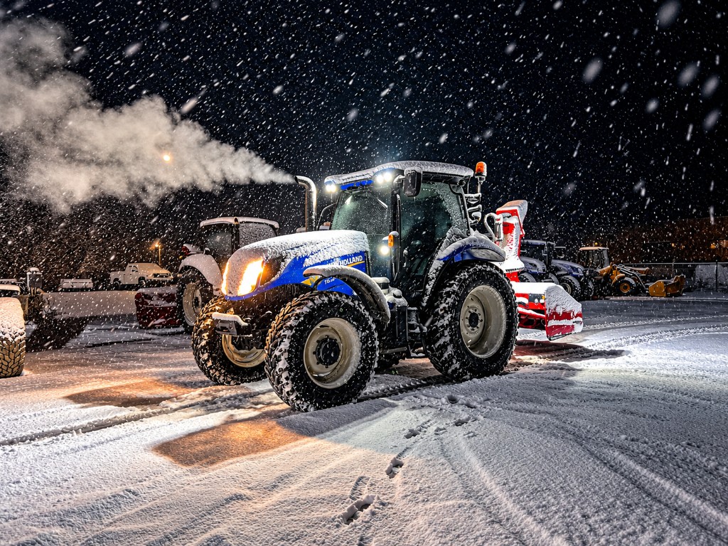 A blue Choreguys New Holland tractor with a red snow blower in the yard at night during a heavy snowfall