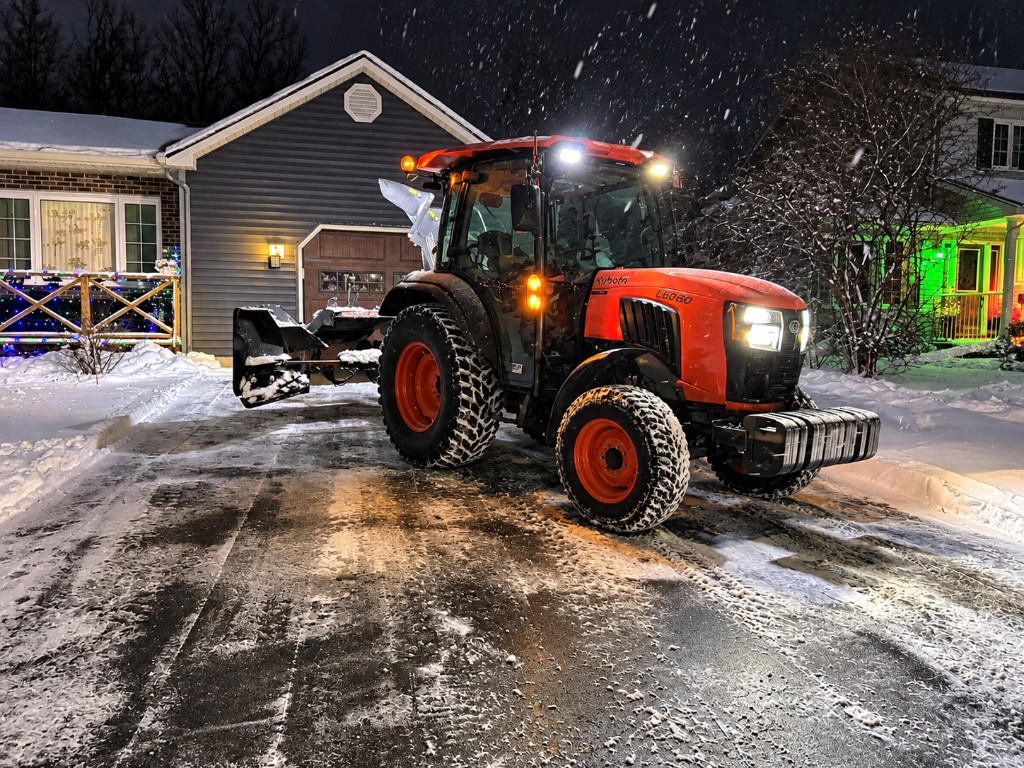 An orange Kubota L6060 tractor with a snow blower parked in a residential driveway at night, snow falling and holiday lights on the houses behind it