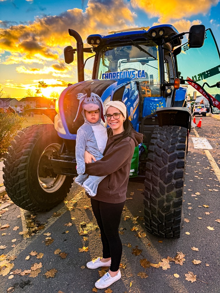 A Choreguys family member in a white beanie and glasses holds a child dressed as Eeyore in front of a blue New Holland tractor at sunset